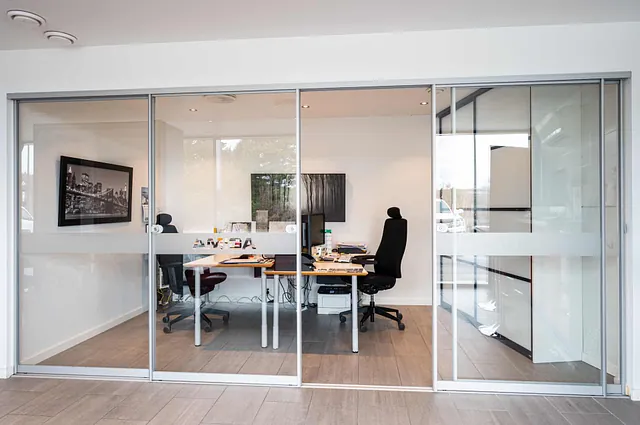 Lyst kontor med glasskyvedører, skrivebord og dataskjermer innenfor.