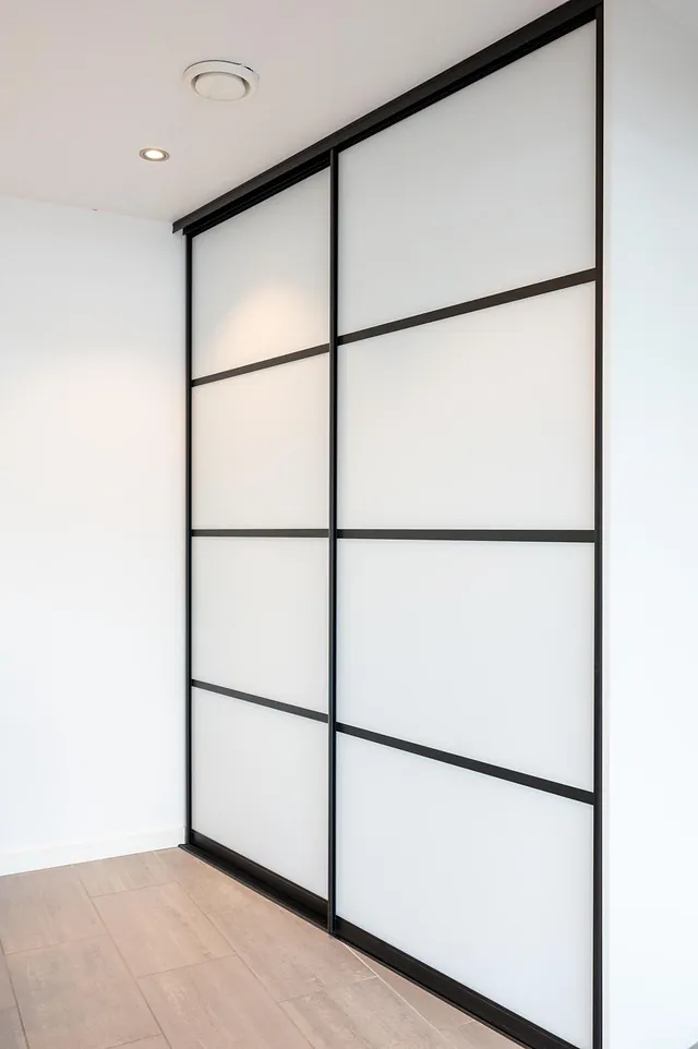 Hvit skyvedørsgarderobe med svarte rammer i rom med lyse vegger og tregulv.