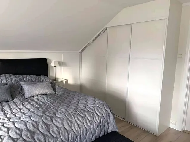 Hvitt soverom med tilpasset garderobe under skråtak og grått sengeteppe.